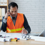 An Asian male construction executive is sitting at his desk in an office, wearing an orange safety vest and a suit. He is talking on the phone while writing on construction blueprints. The desk has a laptop, a calculator, a hard hat, and several tools and documents.