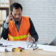 An Asian male construction executive is sitting at his desk in an office, wearing an orange safety vest and a suit. He is talking on the phone while writing on construction blueprints. The desk has a laptop, a calculator, a hard hat, and several tools and documents.