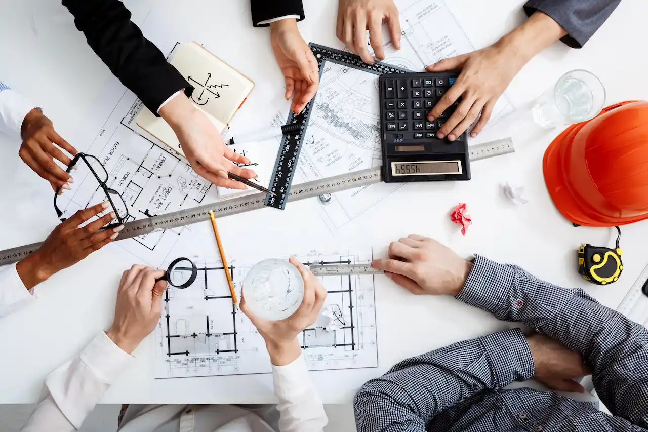 A group of professionals is seen working together at a table with architectural drafts and plans. They are using tools such as a calculator, ruler, magnifying glass, pencil, and a tape measure while reviewing blueprints for a construction project. A safety helmet is also visible on the table.