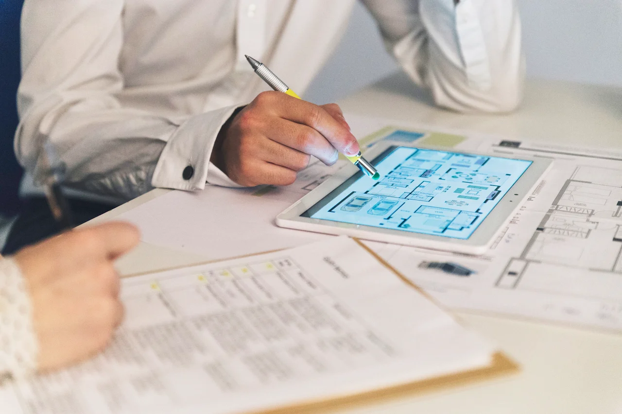 A person is reviewing architectural plans on a tablet while another individual is writing notes on a paper. The plans on the tablet display detailed floor layouts, and they are working together to finalize design details.