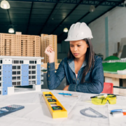 Pensive architect woman in a safety helmet reviewing building model and blueprints in the office.