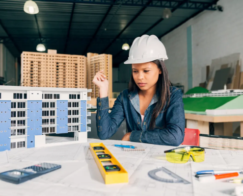 Pensive architect woman in a safety helmet reviewing building model and blueprints in the office.