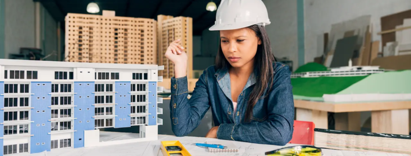 Pensive architect woman in a safety helmet reviewing building model and blueprints in the office.