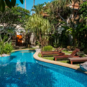 A serene and lush resort-style swimming pool surrounded by tropical plants and lounge chairs. The pool features clear blue water and is nestled within a garden with vibrant greenery, providing a relaxing and peaceful atmosphere.