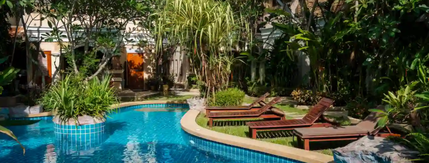 A serene and lush resort-style swimming pool surrounded by tropical plants and lounge chairs. The pool features clear blue water and is nestled within a garden with vibrant greenery, providing a relaxing and peaceful atmosphere.