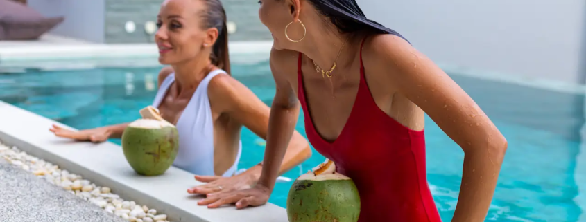 Two women relaxing in a swimming pool holding fresh coconuts by the poolside.